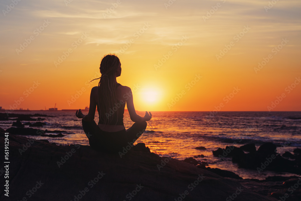 Silhouette young woman sitting in lotus pose, practicing yoga on ocean beach at sunset