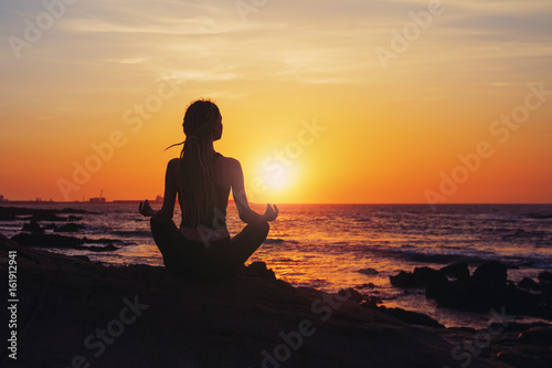 Silhouette young woman sitting in lotus pose, practicing yoga on ocean beach at sunset