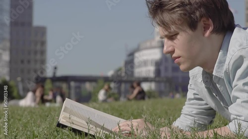 Young Male Lays On The Grass Reading A Book In Slow Motion. Urban Background With Buildings And People On A Sunny Summer Day
