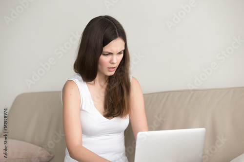 Pretty young woman sitting on sofa and looking with anger on laptop screen. Stressed teen girl working on computer at home. Lady frustrated because of bad news in internet, problems with software