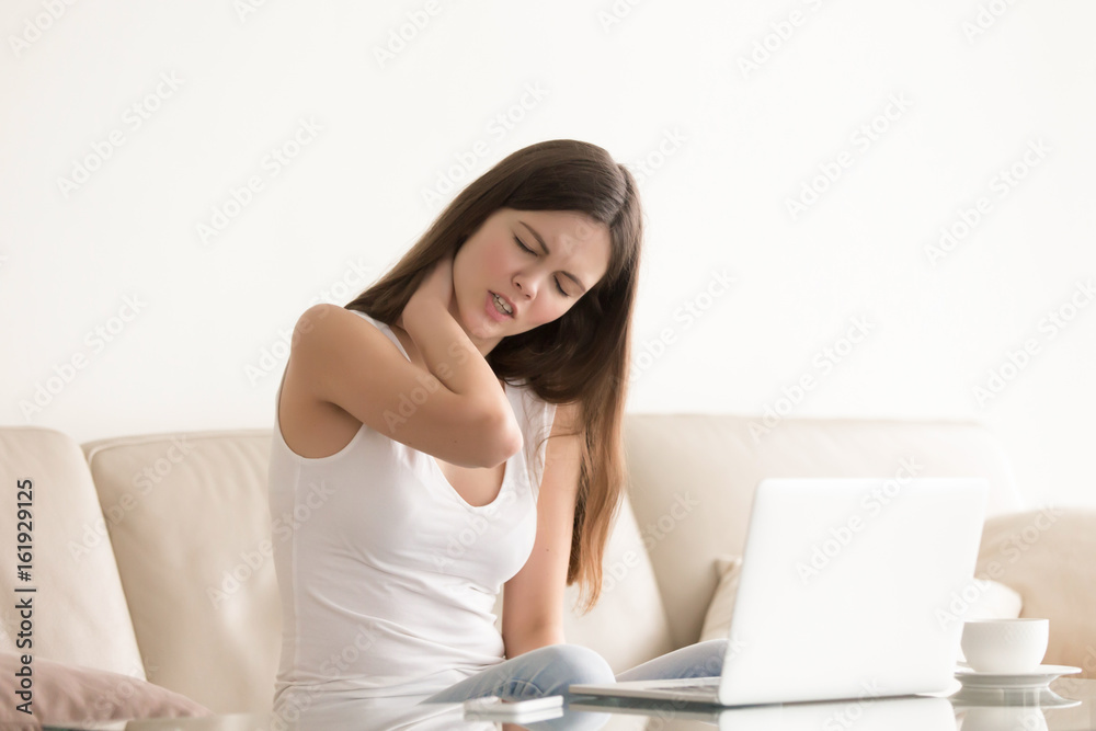 Pretty young woman feels neck pain while sitting on sofa in front of laptop. Tired stressed female suffering from discomfort, massaging tense neck muscles after long hours of sedentary computer work Pretty young woman feels neck pain while sitting on sofa in front of laptop. Tired stressed female suffering from discomfort, massaging tense neck muscles after long hours of sedentary computer work