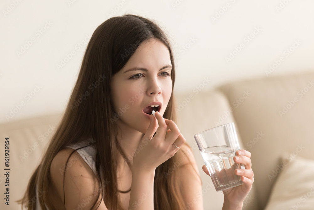 Attractive young woman takes white round pill when feeling sick. Cute teen girl with glass of water in hand drinking medicines, vitamins or supplements. Medical treatment or pharmacological therapy Attractive young woman takes white round pill when feeling sick. Cute teen girl with glass of water in hand drinking medicines, vitamins or supplements. Medical treatment or pharmacological therapy