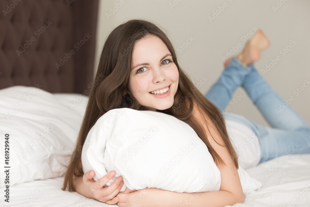 Happy smiling young woman lying on bed with white sheets, hugging a pillow, looking in camera with joy. Relaxed attractive lady feeling positive while resting in luxury hotel room or bedroom. Portrait Happy smiling young woman lying on bed with white sheets, hugging a pillow, looking in camera with joy. Relaxed attractive lady feeling positive while resting in luxury hotel room or bedroom. Portrait
