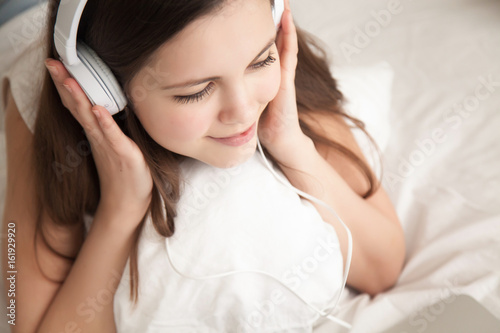 Close up portrait of charming young woman in headphones listening pleasant music while lying in bed with laptop. Attractive teen girl enjoys internet video, watching online movie, studying distantly