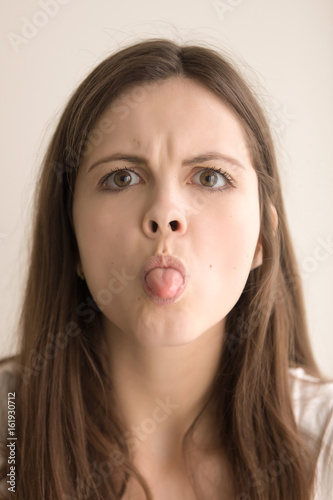 Headshot portrait of woman with bad manners showing tongue. Teen with sticking out tongue and gloomy facial expression looking at camera with wrath. Offended lady feeling angry. Close up. Front view