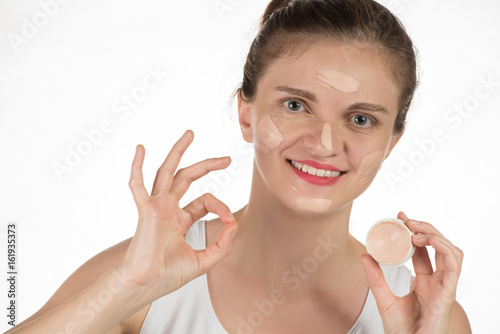 Young brunette woman applying makeup on skin foundation and smiling on white background