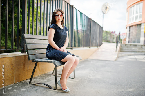 Brunette girl at black dress on sunglasses sitting on bench and posing at street of city.