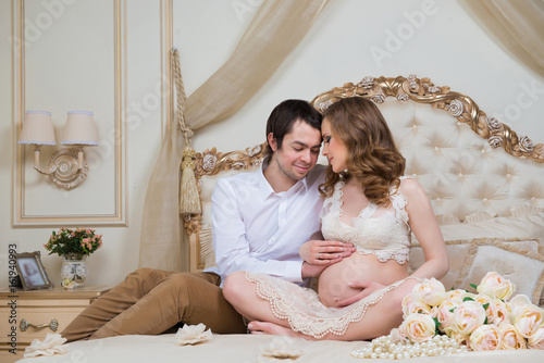Beautiful couple, pregnant young woman and man, hugging lovingly sitting on the bed, in a home interior.