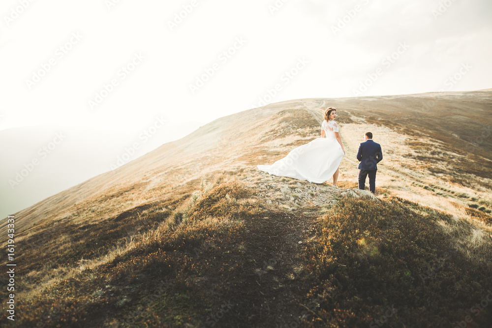 Young newly wed couple, bride and groom kissing, hugging on perfect view of mountains, blue sky