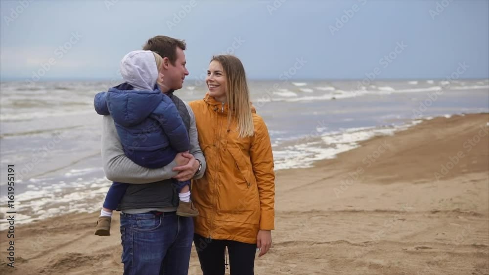 A young married couple is standing on a sandy coast, an adult mother is holding a son in her arms, her middle-aged husband is standing next to him and lovingly looks at his wife A young married couple is standing on a sandy coast, an adult mother is holding a son in her arms, her middle-aged husband is standing next to him and lovingly looks at his wife