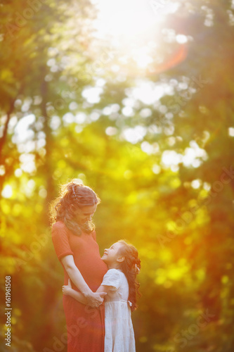 The girl hugs her pregnant mother at the edge of the forest. Woman and daughter are looking at each other in the evening haze and the golden rays of sunset