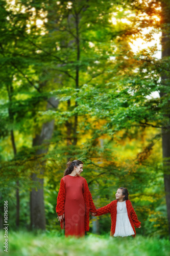 A pregnant woman and her eldest daughter are walking alone along the lush green spring forest, holding each other's hands.