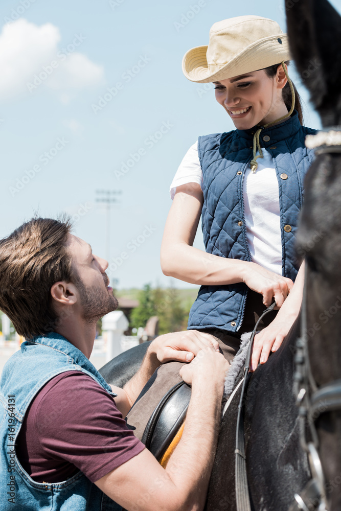 Handsome young man looking at smiling woman sitting on horseback - obrazy, fototapety, plakaty 