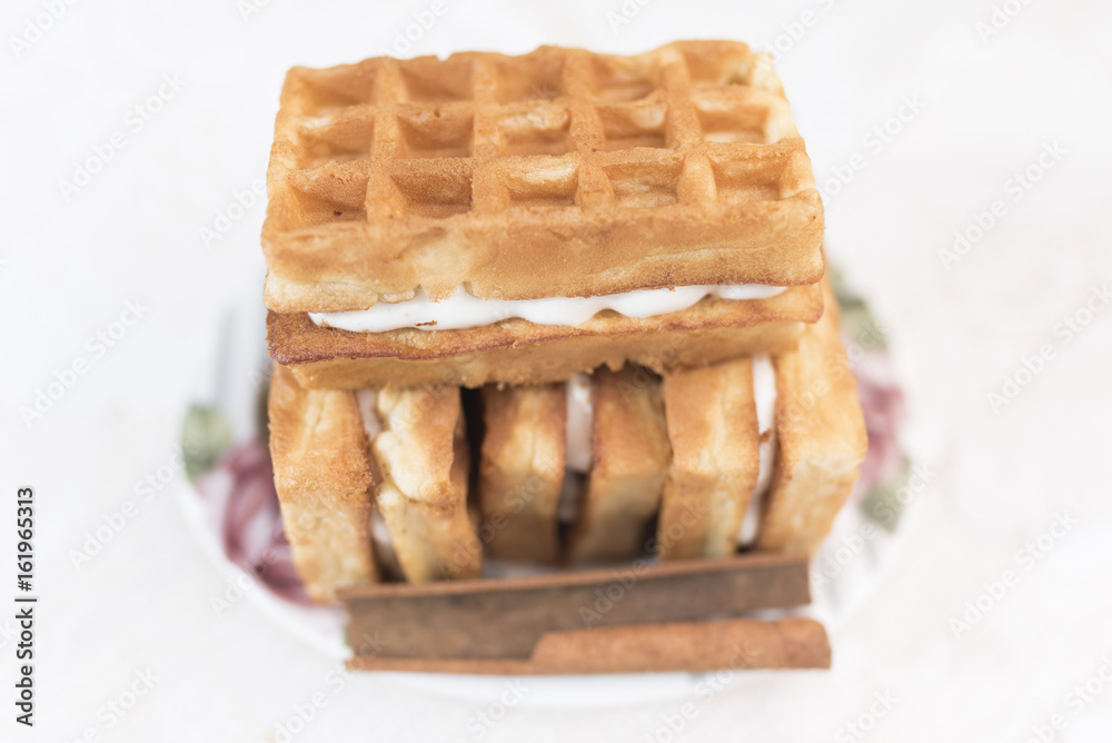 Viennese waffles with cream filling and one stick of cinnamon lie on a white saucer with a floral pattern in the defocus on a white background Viennese waffles with cream filling and one stick of cinnamon lie on a white saucer with a floral pattern in the defocus on a white background