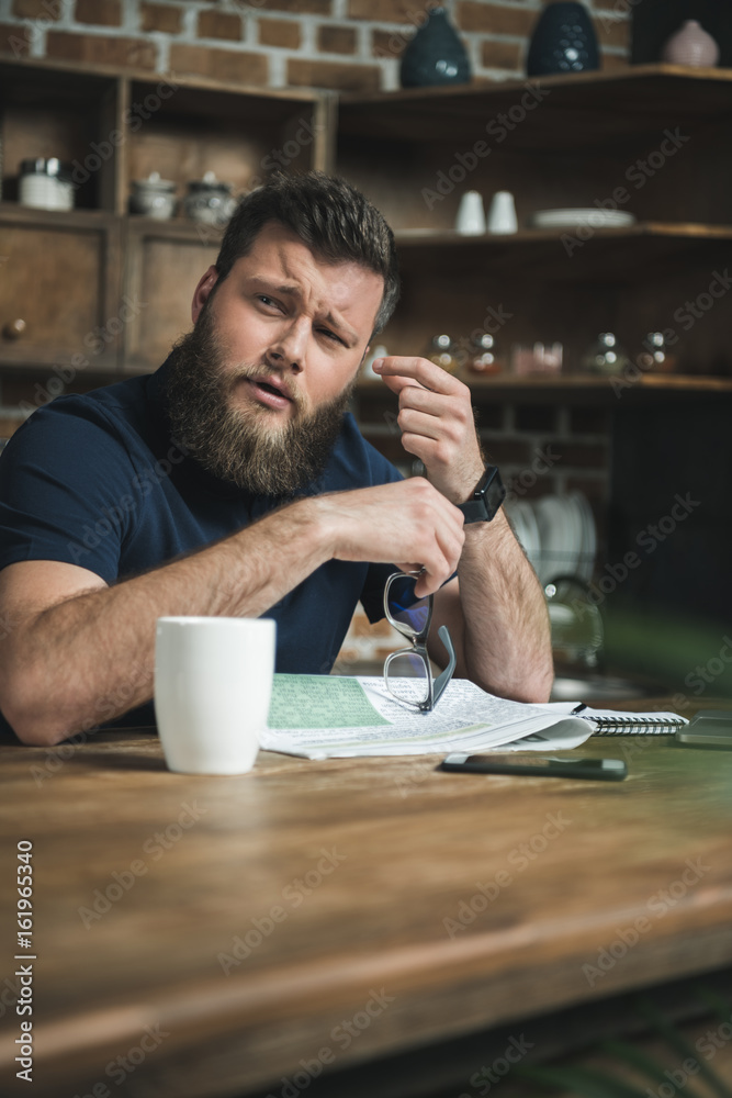 portrait of tired man with eyeglasses in hand looking aside while sitting at table