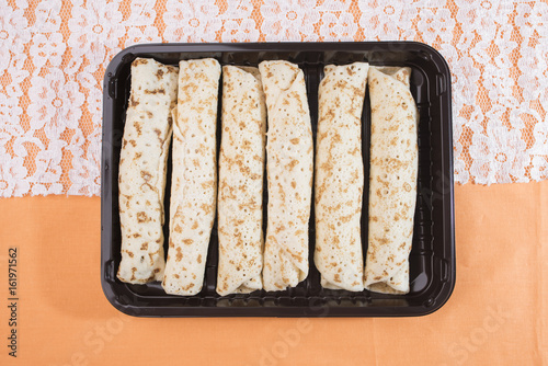 Pancakes rolls with a filling lie in a dark brown tray on a white lace and a delicate peach-orange background