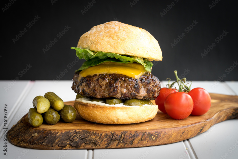 large thick grilled beef minced burger patty with toasted bun melted cheddar cheese green fresh salad cherry tomato and pickle cucumber on rustic wooden board and white table