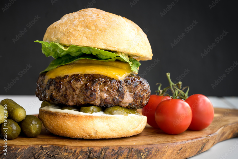 large thick grilled beef minced burger patty with toasted bun melted cheddar cheese green fresh salad cherry tomato and pickle cucumber on rustic wooden board and white table