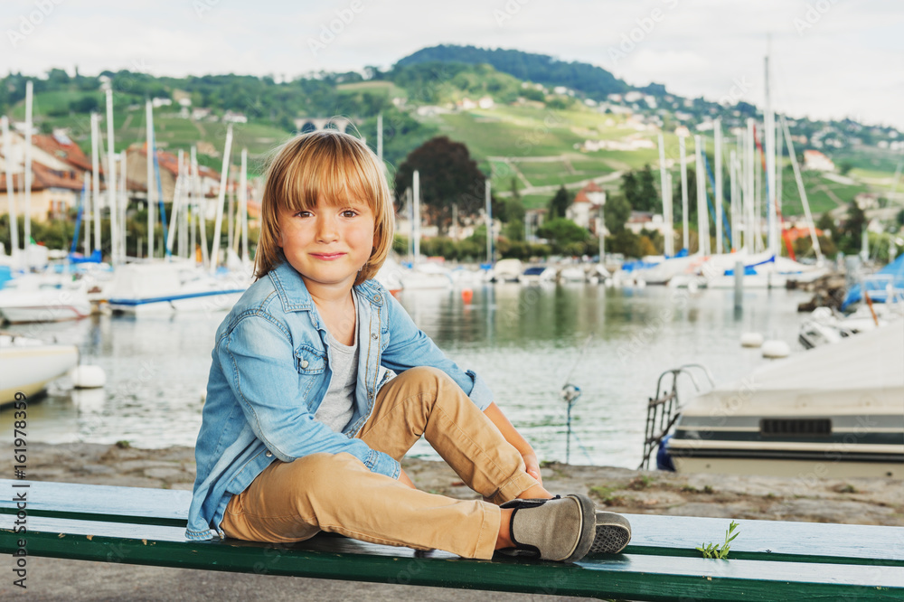 Outdoor portrait of adorable little boy resting on the bench in a beautiful small port on Lake Geneva, Switzerland