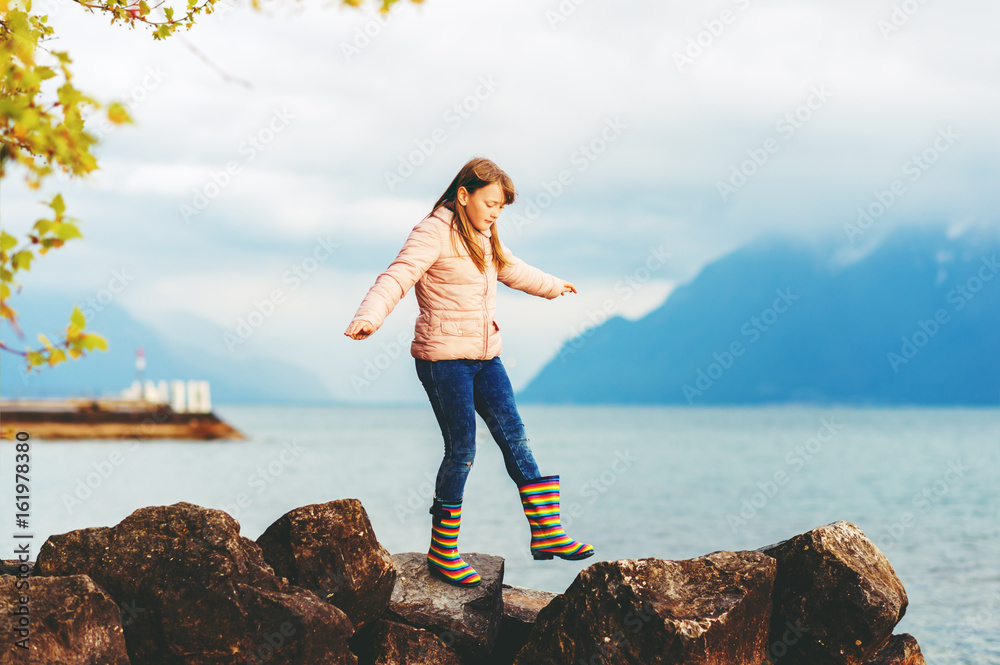 Adorable kid girl playing by the lake on a very cloudy day, wearing warm jacket and rain boots