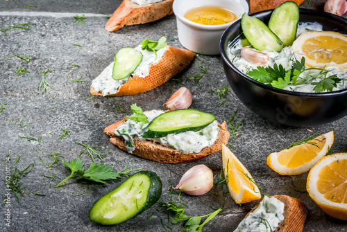 Traditional Caucasian and Greek food. Sauce tzatziki with ingredients - cucumber, lemon, parsley, dill, garlic. On a dark stone table. With sandwiches and baguette. Copy space