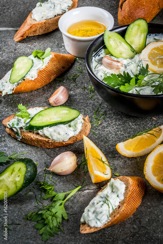 Traditional Caucasian and Greek food. Sauce tzatziki with ingredients - cucumber, lemon, parsley, dill, garlic. On a dark stone table. With sandwiches and baguette.