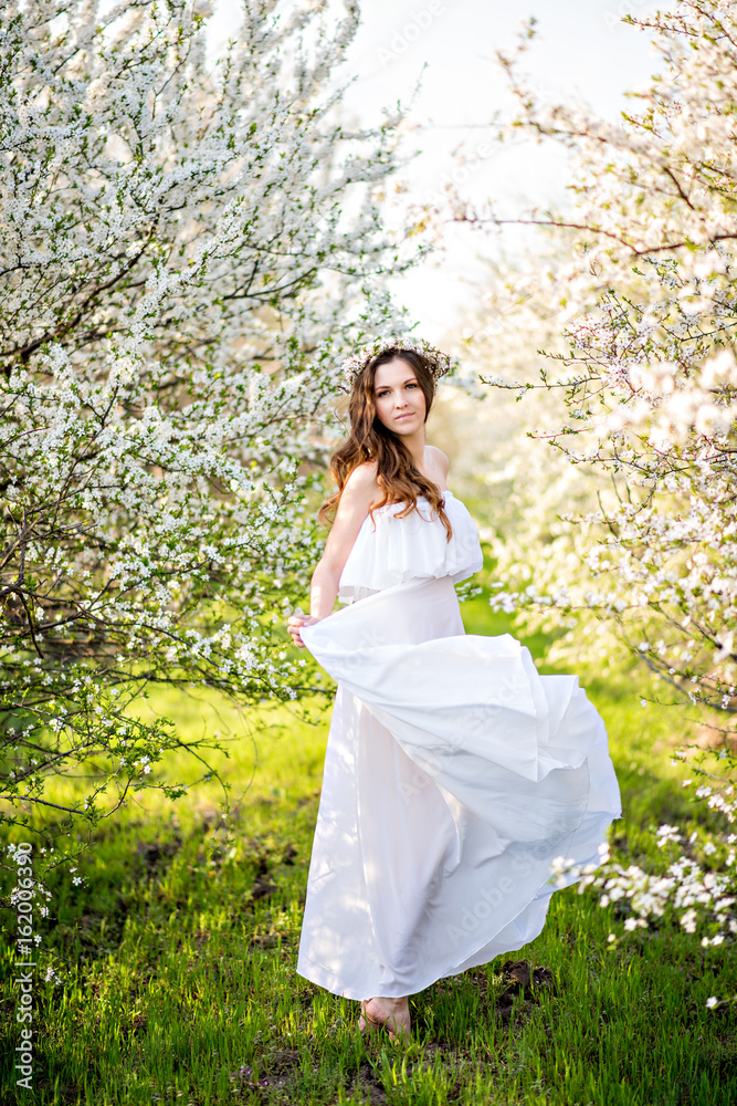 Beautiful young girl in white dress and wreath of flowers on head in blooming gardens Beautiful young girl in white dress and wreath of flowers on head in blooming gardens