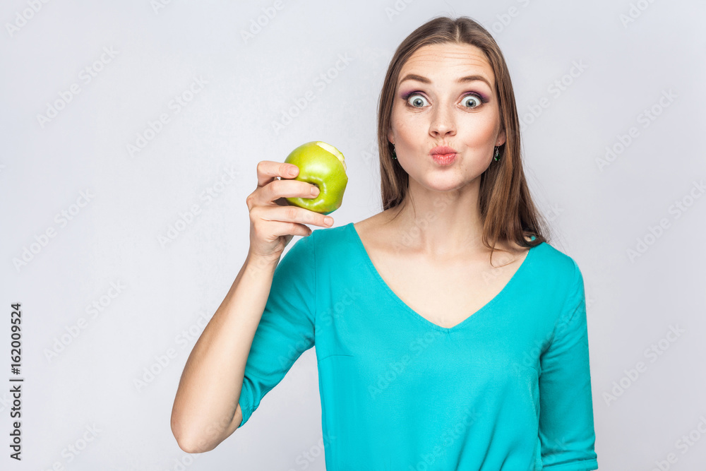 Young beautiful woman with freckles and green dress holding and eating apple and looking at camera with big surprised eyes. studio shot, isolated on light gray background. Young beautiful woman with freckles and green dress holding and eating apple and looking at camera with big surprised eyes. studio shot, isolated on light gray background.