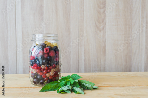 Red, white, black currant, red and black raspberries, white strawberries and mint leaves in glass jar on wooden table as ingredient to healthy cocktail, beverage, yogurt, smoothie