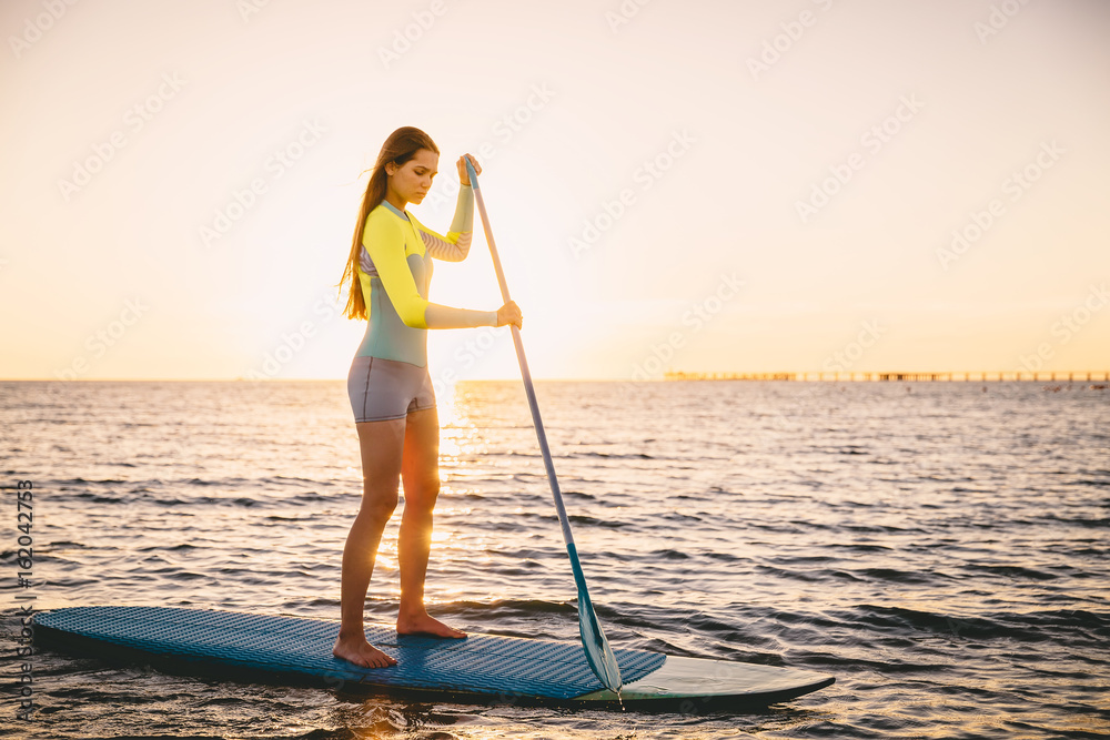 Perfect slim woman stand up paddle surfing in ocean with beautiful sunset colors