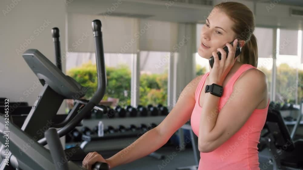 Fit active sportive woman doing exercises on velosimulator. Using her smartphone, talking to friend Fit active sportive woman doing exercises on velosimulator. Using her smartphone, talking to friend