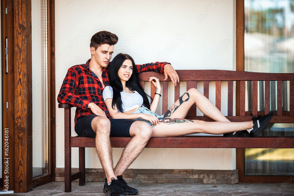 Two hipster models young man and woman with tattoo sitting on the bench with a wistful smile, looking into the distance. Beautiful young couple during a romantic date, urban style Two hipster models young man and woman with tattoo sitting on the bench with a wistful smile, looking into the distance. Beautiful young couple during a romantic date, urban style