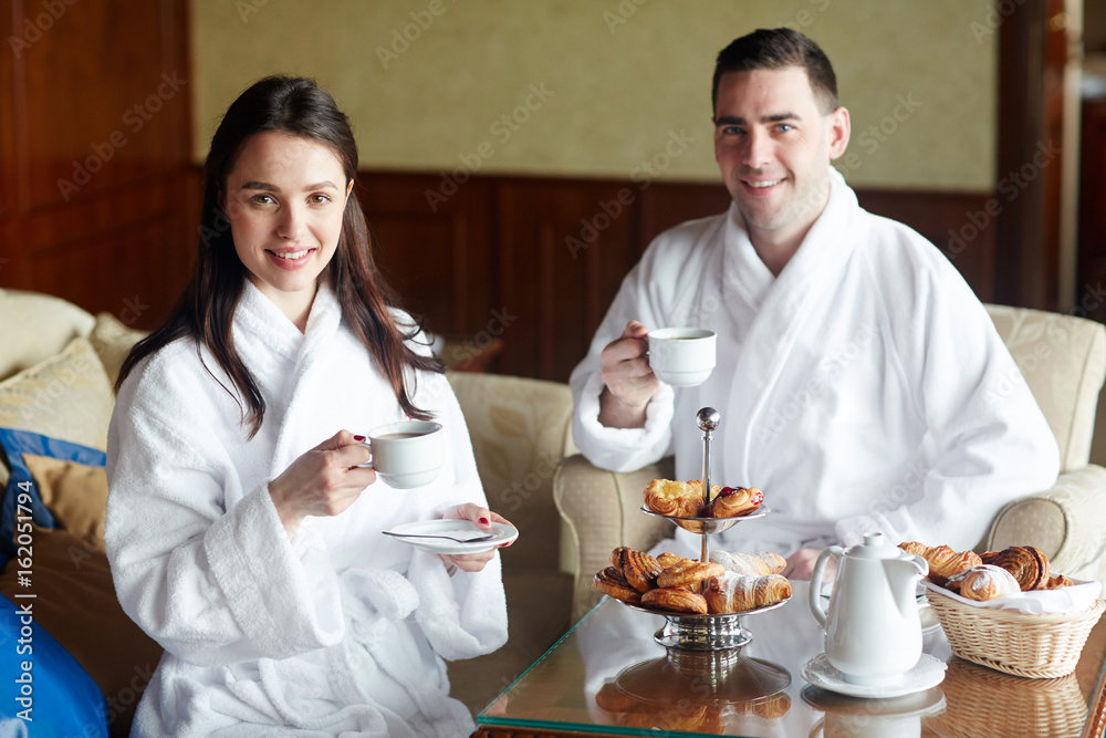 Young couple in bathrobes having coffee with buns in hotel room Young couple in bathrobes having coffee with buns in hotel room