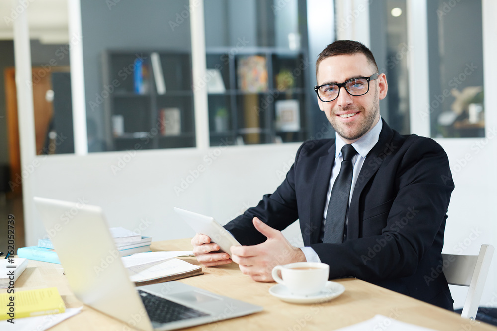 Happy young economist with gadget sitting by his workplace