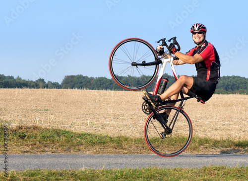 Fototapeta Naklejka Na Ścianę i Meble -  Cheerful cyclist riding on the rear wheel. Biker balancing while driving on a road bike. Risky ride on one wheel.