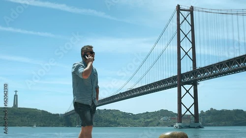 Handsome man walking next to the river while chatting on cellphone, steadycam shot
 photo