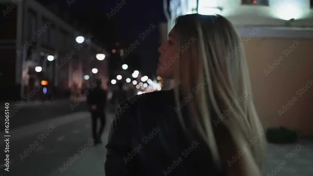 Beautiful young woman against night shined city. The blonde is walking around the night city. Shooting from behind. Beautiful young woman against night shined city. The blonde is walking around the night city. Shooting from behind.