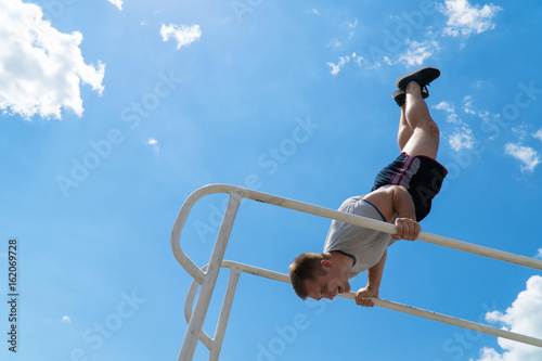 The guy is standing on his hands on the uneven bars. Workout outdoors. Blue sky on blackground