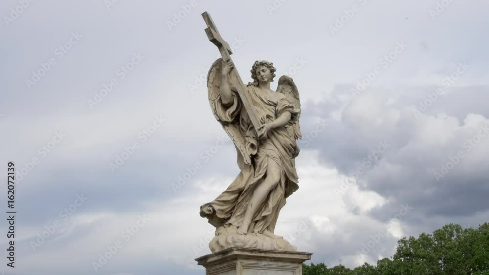 The Angel with the Cross on the Bridge of Hadrian (Ponte Sant’Angelo) in Rome, Italy. Summertime, June. Dramatic clouds in the background. The Angel with the Cross on the Bridge of Hadrian (Ponte Sant’Angelo) in Rome, Italy. Summertime, June. Dramatic clouds in the background.