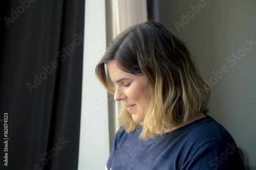 Cute lovely shy smiling young woman standing near the window