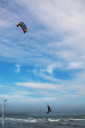  Two kiters are on the waves, one of them is jumping with bright colorful kite against blue sky with white clouds 