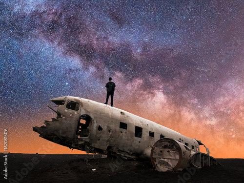 Silhouette of Man Standing on Crashed Airplane Gazes Deeply Into the Milky Way Galaxy on a Clear Starry Night Sky - Horizontal