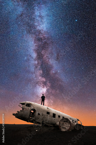 Silhouette of Man Standing on Crashed Airplane Gazes Deeply Into the Milky Way Galaxy on a Clear Starry Night Sky - Vertical