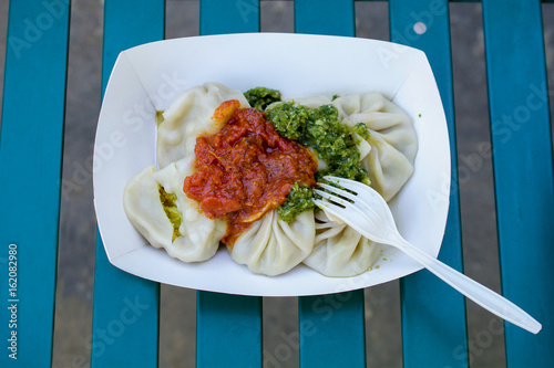 Closeup on Vegie indian momos  - Steamed dumplings with tomato and cilantro sauce.- served in a carry-out box at a farmers market, on a park’s bench , with a plastic fork