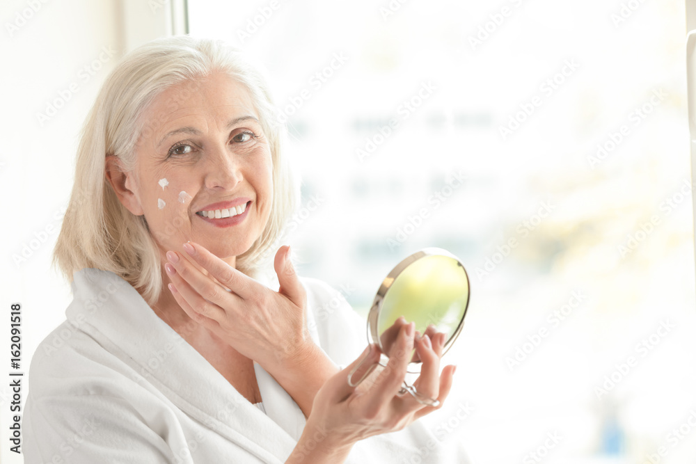 Beautiful elderly woman holding mirror and applying face cream at home Beautiful elderly woman holding mirror and applying face cream at home