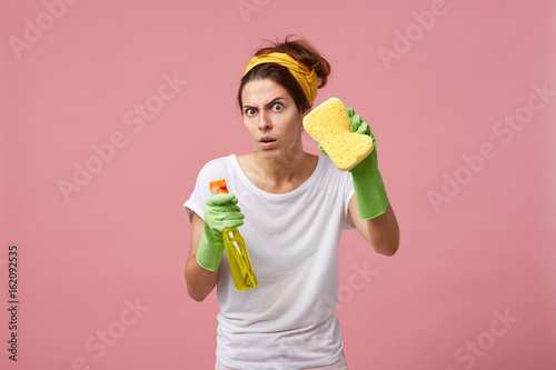 Emotional cute female posing in green rubber gloves, equipped with yellow sponge and spray detergent, ready for tidying up and cleaning house, looking at camera with funny expression on her face