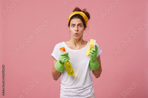 Studio portrait of stressed terrified young Caucasian woman dressed casually feeling panic while has to clean up rooms fast before guests come, holding cleanser spray and sponge in her hands