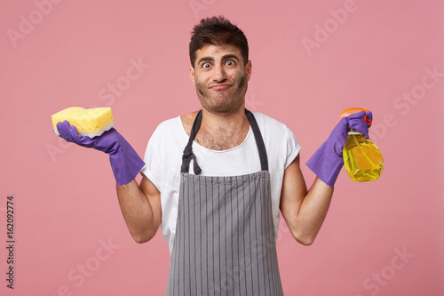 Dirty man with stylish hairdo and bristle holding sponge and detergent wearing casual white T-shirt, apron and gloves being tired after housework helping his wife to clean house. Domestic work