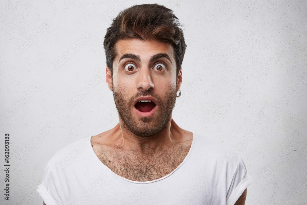 Portrait of stylish bearded guy having trendy hairstyle wearing earring and white T-shirt looking with his eyes popped out and opened mouth having shock and scared look. People and emotions concept Portrait of stylish bearded guy having trendy hairstyle wearing earring and white T-shirt looking with his eyes popped out and opened mouth having shock and scared look. People and emotions concept