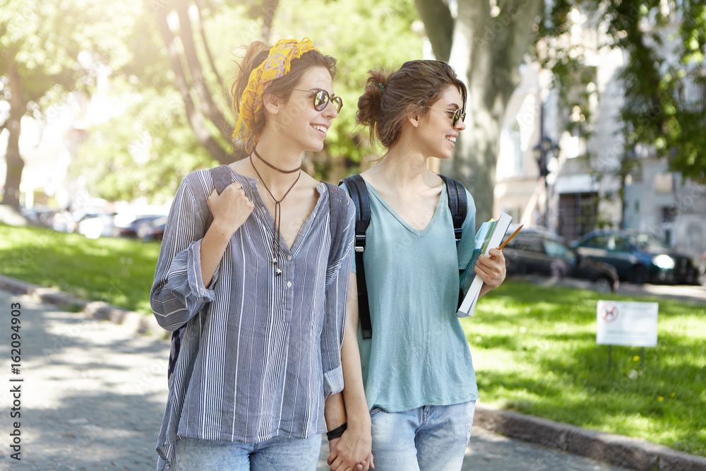 Cheerful students lesbians walking togehter across street holding hands together looking with smile aside. Homosexual women in summer clothes and sunglasses having fun together admiring nature